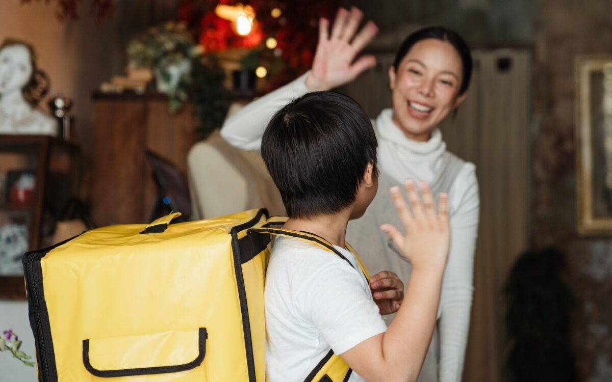 Happy ethnic female cafe worker in apron wishing farewell to boy with food delivery backpack helping in family business