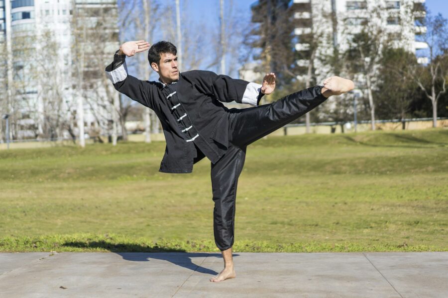 Young athletic martial arts fighter practicing kicks in a public park wearing kung fu uniform.